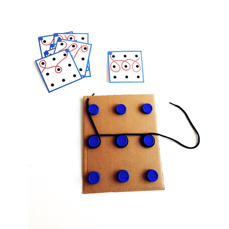 Op de afbeelding: Een kartonnen bord met blauwe plastic flesdoppen eraan bevestigd. Een zwart touwtje is aan de doppen vastgemaakt. Er zijn ook verschillende witte kaartjes met zwarte stippen en rode lijnen.