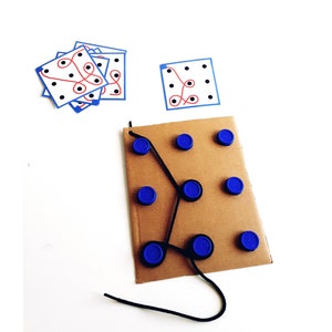Op de afbeelding: Een kartonnen spelbord met blauwe dopjes en een zwart touwtje. Het touwtje is door de dopjes geregen en vormt een labyrintpatroon. Er zijn ook verschillende bedrukte kaartjes met zwarte stippen en rode en blauwe lijnen.