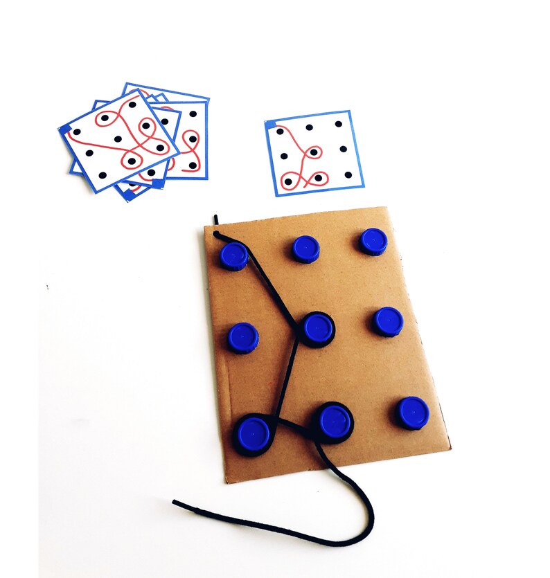 Op de afbeelding: Een kartonnen spelbord met blauwe dopjes en een zwart touwtje. Het touwtje is door de dopjes geregen en vormt een labyrintpatroon. Er zijn ook verschillende bedrukte kaartjes met zwarte stippen en rode en blauwe lijnen.