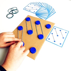 May include: A child's hands interacting with a DIY educational toy. The toy features a cardboard base with blue bottle caps and rubber bands, alongside pattern cards with blue dot designs. Green rubber bands and additional blue bottle caps are also visible.