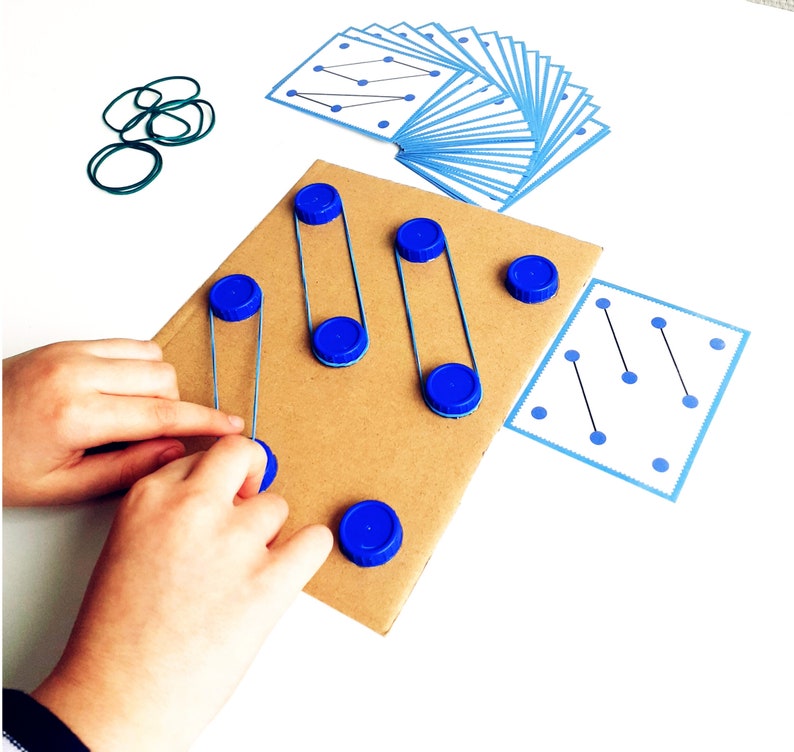 May include: A child's hands interacting with a DIY educational toy. The toy features a cardboard base with blue bottle caps and rubber bands, alongside pattern cards with blue dot designs. Green rubber bands and additional blue bottle caps are also visible.