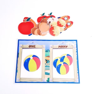 May include: A wooden frame with two sections labeled "ONE" and "MANY".  The left section has one beach ball, and the right section has three beach balls.  There are various colorful cutouts above the frame, including apples, acorns, and lollipops.