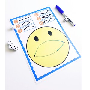 May include: A yellow smiley face drawing with a green mouth and black eyes on a white paper with a blue border. The paper has numbered line drawings and is next to dice and a blue marker.