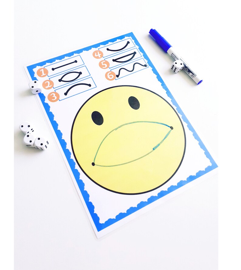 May include: A yellow smiley face drawing with a green mouth and black eyes on a white paper with a blue border. The paper has numbered line drawings and is next to dice and a blue marker.