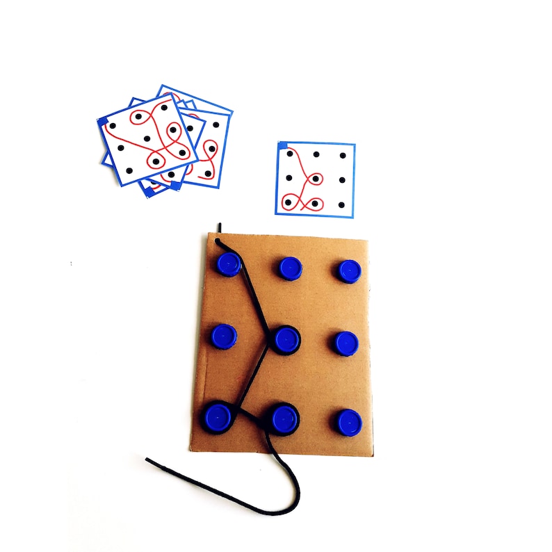 Op de afbeelding: Een kartonnen spelbord met blauwe plastic cirkels en een zwarte draad eraan bevestigd. Het bord heeft een rood en blauw patroon. Er zijn ook verschillende witte kaarten met rode en blauwe patronen.