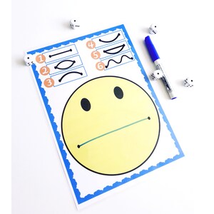 May include: A white paper with a yellow smiley face and a blue border. The paper has numbered boxes with different line shapes. A blue marker and several dice are next to the paper.