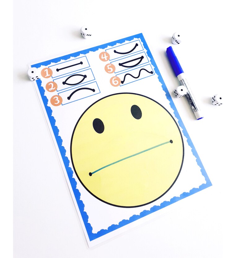 May include: A white paper with a yellow smiley face and a blue border. The paper has numbered boxes with different line shapes. A blue marker and several dice are next to the paper.
