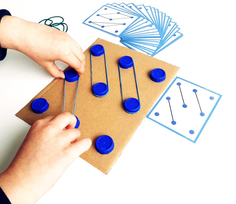 May include: A child's hands interacting with a homemade educational toy. The toy features a cardboard base with blue bottle caps and wire, along with pattern cards. The child is manipulating the blue caps and wire.