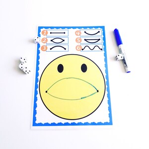 May include: A yellow smiley face drawing with a green curved line for a mouth, and black eyes. The drawing is on a white paper with a blue border. There are six line drawings above the smiley face. A blue marker and dice are on the white surface.