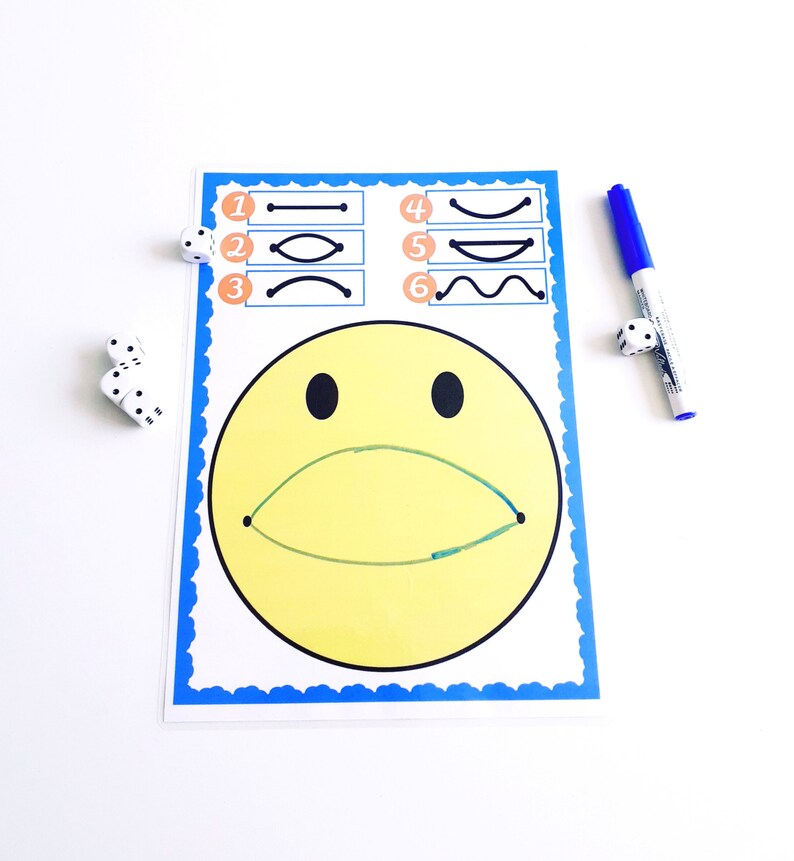 May include: A yellow smiley face drawing with a green curved line for a mouth, and black eyes. The drawing is on a white paper with a blue border. There are six line drawings above the smiley face. A blue marker and dice are on the white surface.