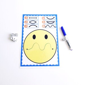 May include: A learning activity sheet with a large yellow smiley face and a green line. The sheet has numbered line patterns and is accompanied by dice and a blue marker. The sheet has a blue border.