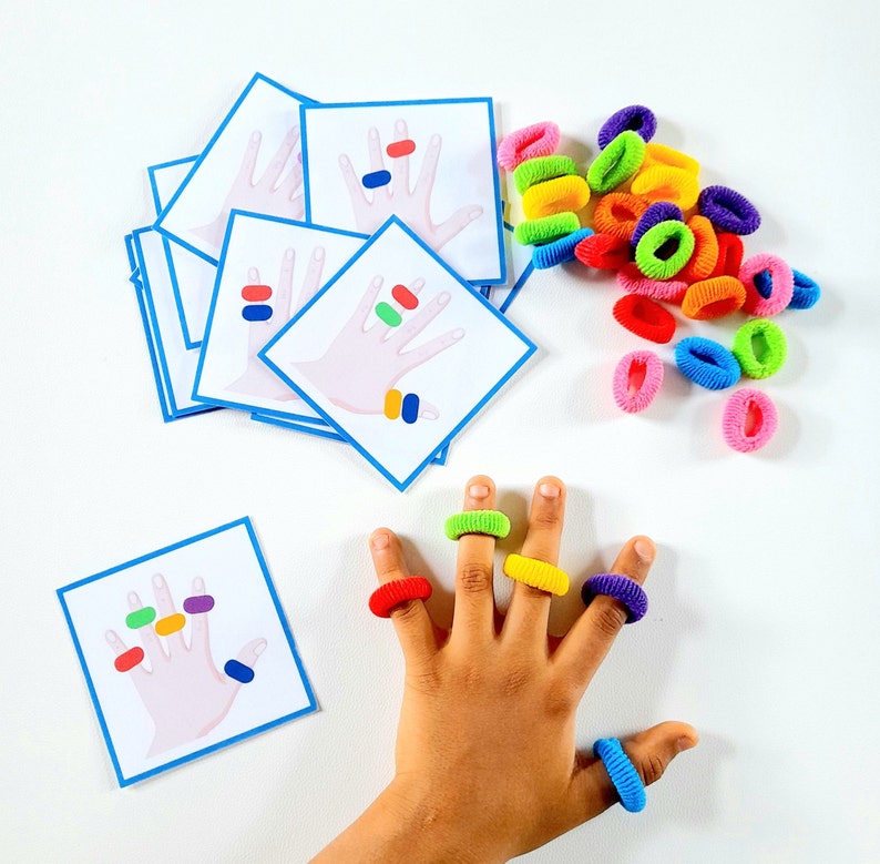 K&ouml;nnte beinhalten: Ein Satz von f&uuml;nf bunten Karten mit Bildern von H&auml;nden und verschiedenen farbigen Punkten an jedem Finger. Die Karten sind f&uuml;r eine Fingerz&auml;hl-Aktivit&auml;t konzipiert. Es gibt auch bunte Gummib&auml;nder im Bild.