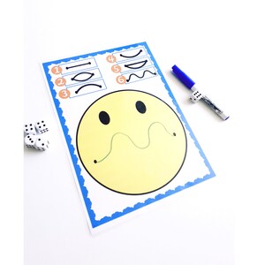 May include: A white paper with a blue scalloped border features a yellow smiley face and line tracing activities. A blue marker and dice are next to the paper. The image is on a white surface.