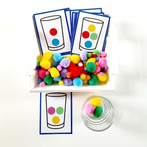 Peut inclure: Un ensemble de pompons colorés dans un plateau blanc avec une bordure bleue. Il y a trois cartes avec des images de verres contenant des points de différentes couleurs. Les cartes sont également bordées de bleu. Il y a un pot en verre transparent avec quelques pompons à l'intérieur.