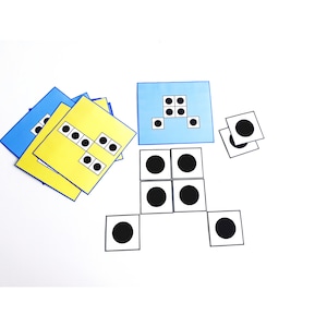May include: A collection of colorful square cards with black dots arranged in various patterns. Some cards are blue or yellow, while others are white with large black circles. The cards are scattered on a white surface, suggesting a game or educational activity.
