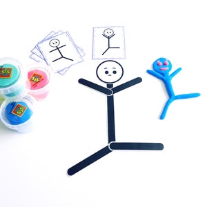 May include: A black and white stick figure with a smiling face, black arms and legs, and a black body. The stick figure is on a white background. There are also three containers of play dough in the image, one green, one pink, and one blue.