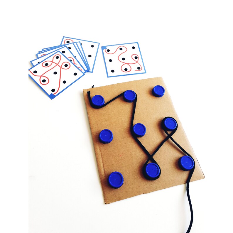 Op de afbeelding: Een kartonnen spelbord met blauwe dopjes en een zwart touwtje. Het touwtje is door de dopjes geregen, waardoor een labyrintpatroon ontstaat. Er zijn verschillende kaarten met rode en zwarte lijnen op een witte achtergrond.