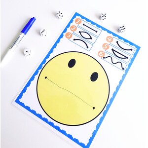 May include: A yellow smiley face drawing with a blue border and a line for a mouth. A blue marker and several dice are scattered around the drawing. The drawing also has lines to trace.