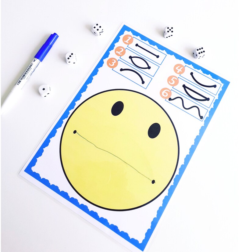 May include: A yellow smiley face drawing with a blue border and a line for a mouth. A blue marker and several dice are scattered around the drawing. The drawing also has lines to trace.