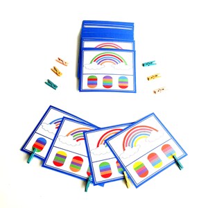 Puede incluir: Un conjunto de tarjetas de aprendizaje con temática de arcoíris con un arcoíris, nubes y una paleta de colores de arcoíris. Las tarjetas son azules con bordes blancos. También hay pinzas para la ropa de colores.
