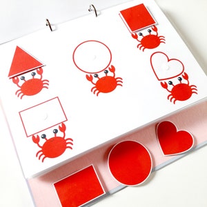 May include: A red, white, and black educational toy for children. The toy features a variety of shapes, including a circle, square, triangle, and heart. Each shape has a red crab holding it up.
