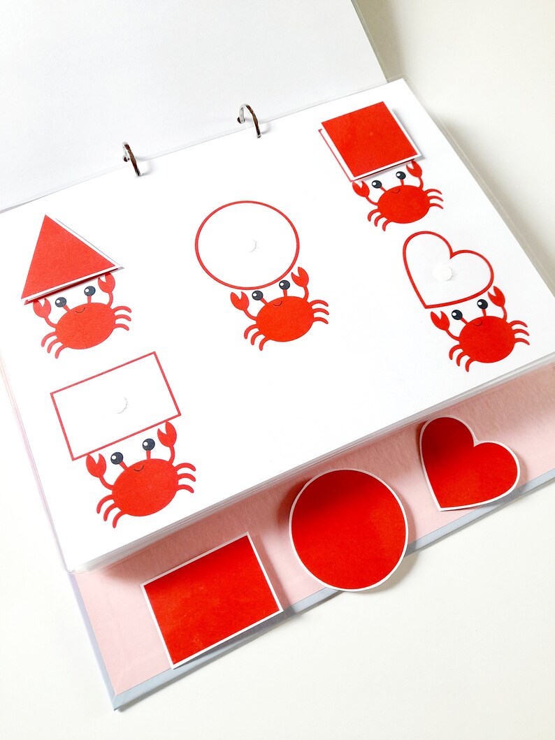May include: A red, white, and black educational toy for children. The toy features a variety of shapes, including a circle, square, triangle, and heart. Each shape has a red crab holding it up.