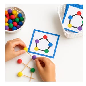 Może przedstawiać: Ręce dziecka montują kolorową zabawkę edukacyjną. Zabawka składa się z małych, kolorowych kulek i wykałaczek, ułożonych tak, aby pasowały do wzorów na kartach. Biały kosz zawiera więcej kulek, a drugi pasujące karty.