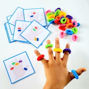 K&ouml;nnte beinhalten: Eine Kinderhand tr&auml;gt an jedem Finger bunte Gummib&auml;nder. Die Hand h&auml;lt eine Karte mit einer Zeichnung einer Hand mit f&uuml;nf Fingern, wobei jeder Finger einen andersfarbigen Punkt hat. Die Karte ist von anderen Karten mit &auml;hnlichen Zeichnungen umgeben.