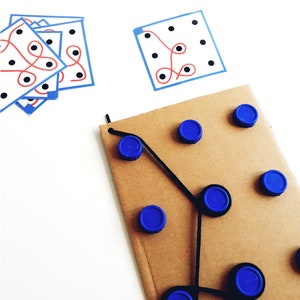 Op de afbeelding: Een bruin kartonnen oppervlak met blauwe dopjes die in een patroon zijn gerangschikt. Zwarte draad verbindt de dopjes en cre&euml;ert een labyrint-achtig ontwerp. Er zijn twee witte kaarten met zwarte stippen en rode en blauwe lijnen op een witte achtergrond.