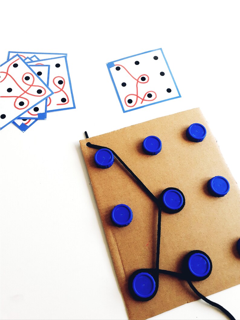 Op de afbeelding: Een bruin kartonnen oppervlak met blauwe dopjes die in een patroon zijn gerangschikt. Zwarte draad verbindt de dopjes en cre&euml;ert een labyrint-achtig ontwerp. Er zijn twee witte kaarten met zwarte stippen en rode en blauwe lijnen op een witte achtergrond.