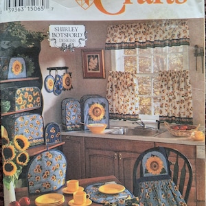 May include: A Simplicity Crafts sewing pattern envelope 3880 by Shirley Botsford Designs featuring illustrations of kitchen textiles including curtains, appliance covers, placemats, and chair covers, all decorated with a sunflower and blue gingham pattern.