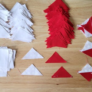 May include: An overhead shot shows various fabric pieces for quilting on a wooden surface. There are stacks of white squares and triangles, along with red triangles and assembled red and white shapes. The pieces are arranged for a quilting project.