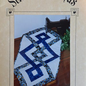 May include: A sewing pattern cover titled Stitchin' Friends Simply Squares #981 featuring a table runner with a geometric blue and white square design, accented by a black fabric border with a blue daisy floral print, displayed near a sewing machine.
