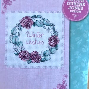 Op de afbeelding: Een kruissteekontwerp met een krans van roze rozen en groene bladeren, met de woorden "Winter wishes" in het midden. Het ontwerp staat op een lichtroze achtergrond met bloemaccenten en een label met de tekst "Exclusive Durene Jones Design."