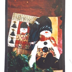 May include: A fabric snowman doll wearing a black knit cap with white cross-stitch detail, a red plaid scarf, and black buttons on its chest. Behind it sits a rectangular fabric sign with the word Snow, black tree appliques, and buttons.