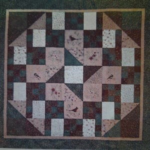 May include: A square quilt with a patchwork design featuring squares, triangles, and rectangles in shades of brown, green, and white. The quilt has a bird and floral pattern. The quilt is bordered with a light brown fabric.