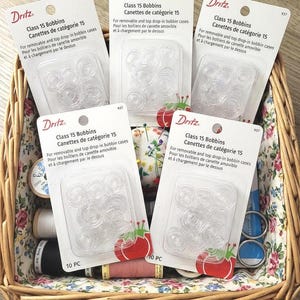 May include: A wicker basket filled with sewing supplies, including multiple packages of Dritz Class 15 Bobbins. Each package contains clear plastic bobbins. The packages have text in English and French. Also visible are spools of thread, a pincushion, and scissors.