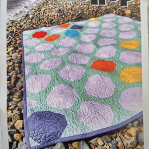 May include: A quilt pattern called "Dollop" from Carolina Patchworks, no. 043. The quilt is a hexagonal pattern with a green background and purple, orange, and yellow accents. The quilt is photographed on a bed of rocks.