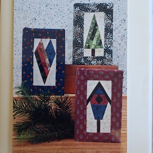 Op de afbeelding: Een patroonboekje getiteld NOVEL COVER-UPS: A QUILTED BOOKCOVER met drie gewatteerde boekomslagen met hart-, boom- en vogelhuisjesontwerpen. De omslag toont de tekst the Plaid Cat La Center, Washington en een blauwe gingham kattenafbeelding.