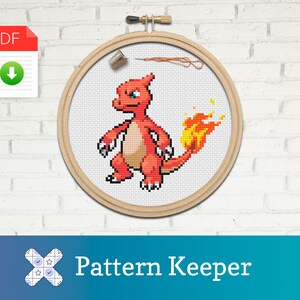 Puede incluir: Un patrón de punto de cruz de un personaje Pokémon rojo y naranja, Charmeleon, en un aro de bordado de madera. El patrón está disponible para descargar en PDF.