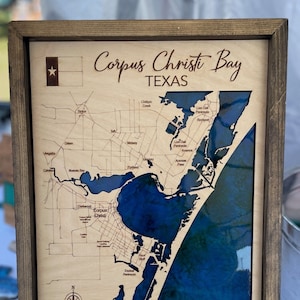 May include: A wooden map of Corpus Christi Bay, Texas, with a blue resin inlay representing the water. The map is framed in a dark wood frame and is displayed on a small easel.