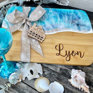 May include: Personalized bamboo cutting board with a blue and white ocean wave design. The board is decorated with a brown ribbon and a wooden tag that says "Sold" with a real estate agent's contact information. The board is also engraved with the name "Lyon".