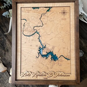 May include: A wooden map of Lake Lyndon B Johnson in Texas, with a blue-green lake and brown land features. The map is framed in a dark brown wood frame.