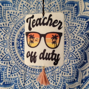 May include: White car air freshener with a black string and a pink tassel. The air freshener has a graphic of sunglasses with palm trees reflected in the lenses. The text "Teacher off duty" is printed on the air freshener.