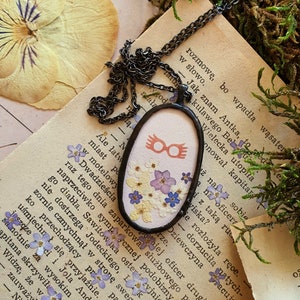 May include: A delicate necklace with a silver chain and a round pendant featuring a pair of red-rimmed glasses and pressed flowers. The pendant is set against a white background.
