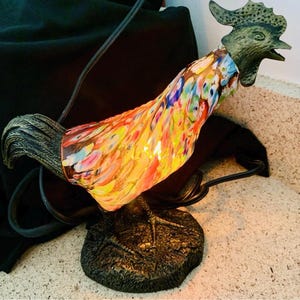 May include: A decorative rooster lamp with a colorful, stained-glass body and a bronze-colored head and base. The lamp's body is a vibrant mix of red, yellow, blue, and green. The lamp is plugged in with a black cord.