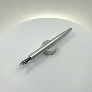 May include: A silver fountain pen with a black nib rests on a small, circular silver stand. The pen has a sleek, modern design and is angled slightly across the stand. The background is a bright, white surface.