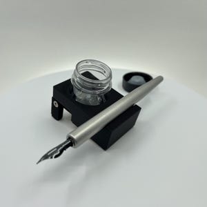 Peut inclure: Un stylo de calligraphie argenté avec une plume noire repose sur un support d'encrier noir. Un pot d'encre en verre transparent est posé au-dessus du support, et un couvercle d'encrier noir est à proximité. L'ensemble est conçu pour la calligraphie et l'écriture.