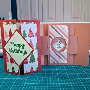 Può includere: Due biglietti di Natale fatti a mano. Un biglietto è aperto, mostrando uno sfondo bianco con alberi di Natale rossi e verdi e il testo "Happy Holidays". L'altro biglietto è chiuso, con un motivo a righe e il testo "to you and Yours."