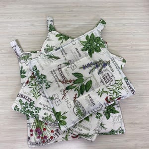 May include: White cotton pot holders with a botanical herb print. The design features various herbs like thyme, parsley, and sage, along with text. Each pot holder has a loop for hanging.